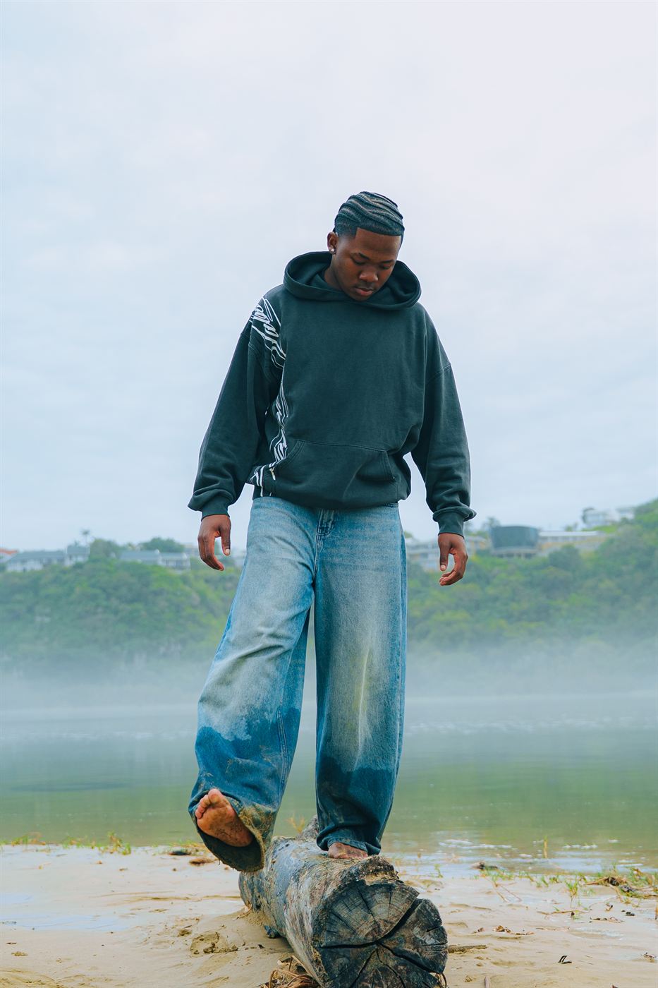 Lifestyle photography — model in a hoodie balancing barefoot on a driftwood log by misty water