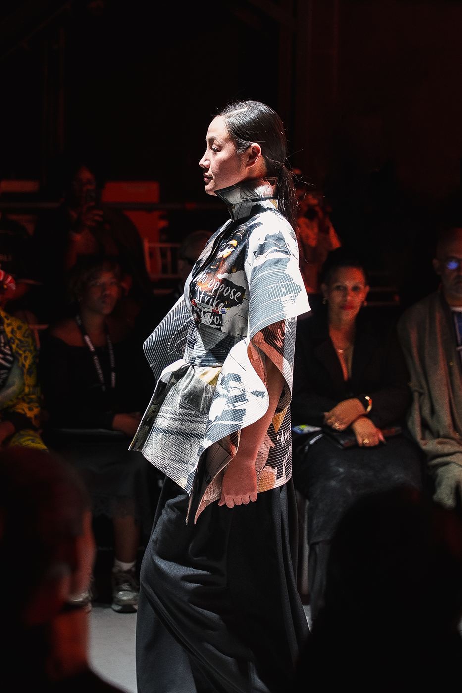 Runway photography — model in a sculptural printed jacket, side profile under stage light