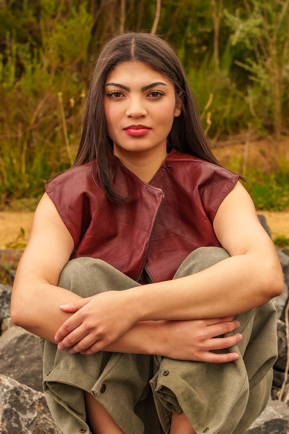 Editorial portrait — model in a burgundy leather vest seated on rocks