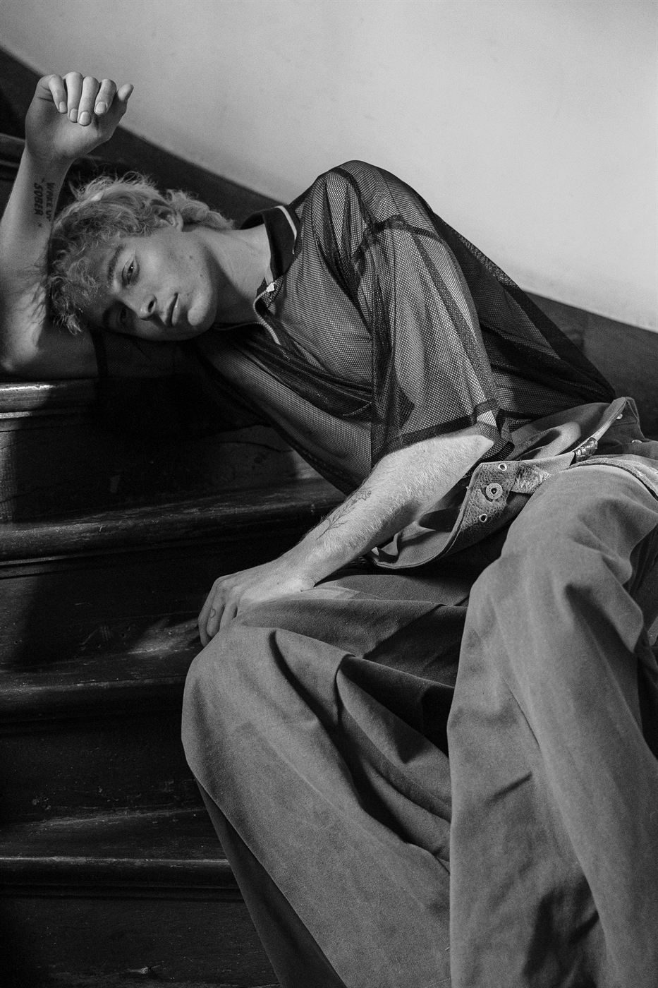 Black and white editorial — model in a mesh shirt reclining on wooden stairs