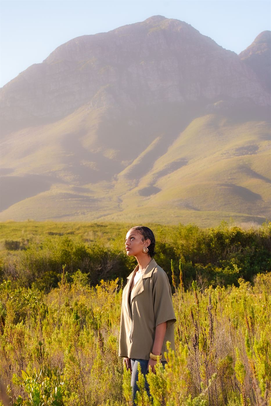 Editorial portrait — model in an olive shirt standing in fynbos below a mountain at golden hour
