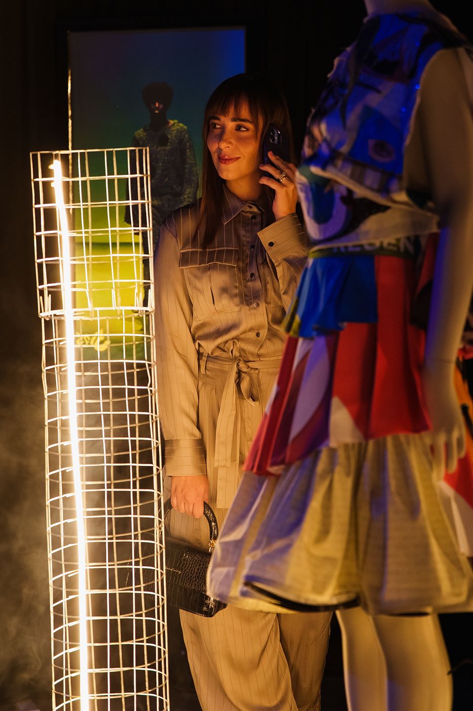 Event photography — guest in a satin jumpsuit on a phone call beside a neon-lit sculpture at fashion week